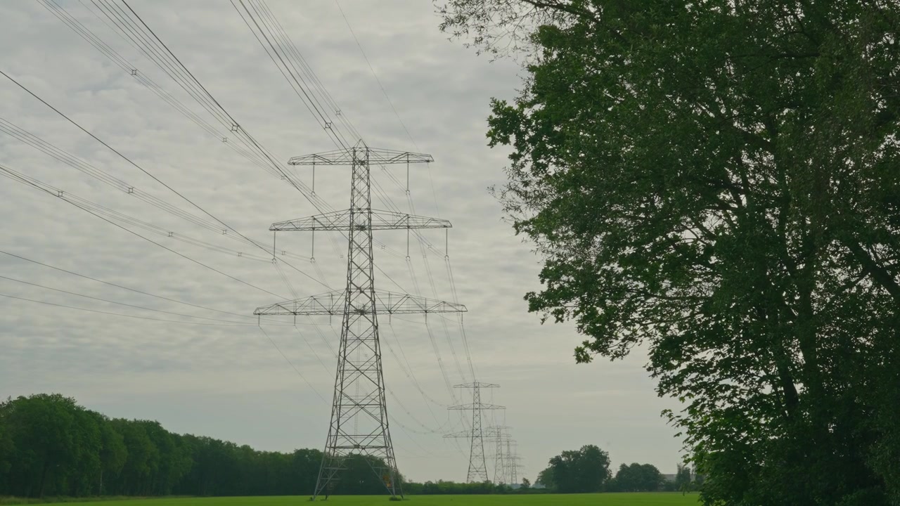 Large electrical transmission towers and power lines stretch across a green agricultural landscape surrounded by trees. A rural scene showing the connection between energy infrastructure and nature.
