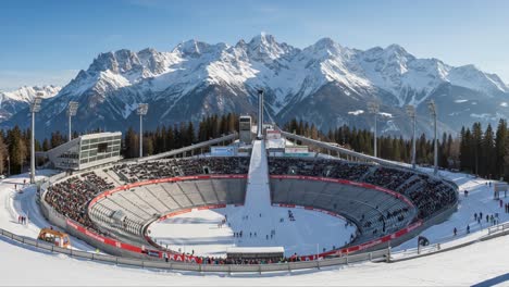 a panoramic view of a winter sports stadium surrounded by majestic snow-capped mountains, filled with spectators enjoying the thrill of the event
