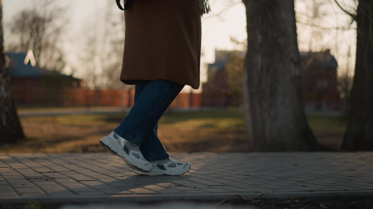 A person wearing a brown coat, jeans, and white shoes is shown walking slowly through a park at sunset. The focus is on their legs, emphasizing their slow, sorrowful steps