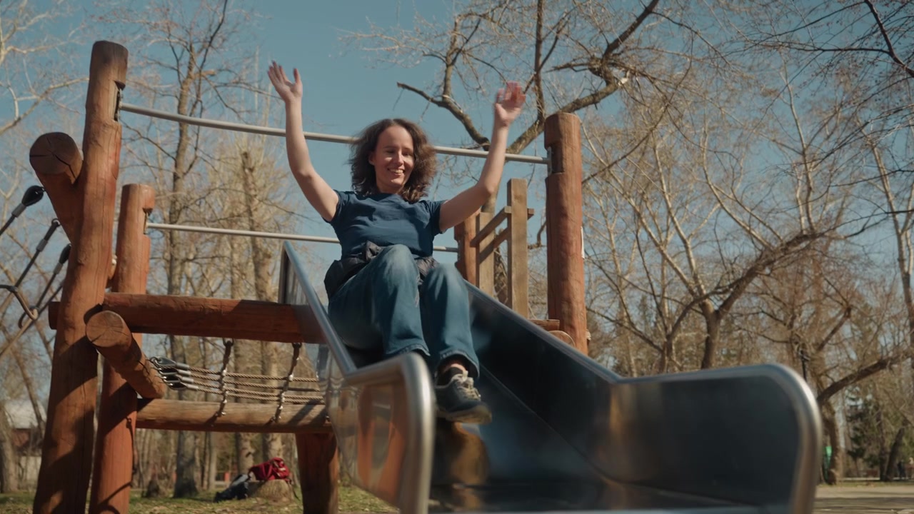 Una donna che indossa una maglietta blu e jeans scende da uno scivolo da parco giochi all'aperto, tenendosi alla barra in cima prima di sedersi per godersi la discesa. La scena presenta strutture di gioco in legno con barre di metallo.
