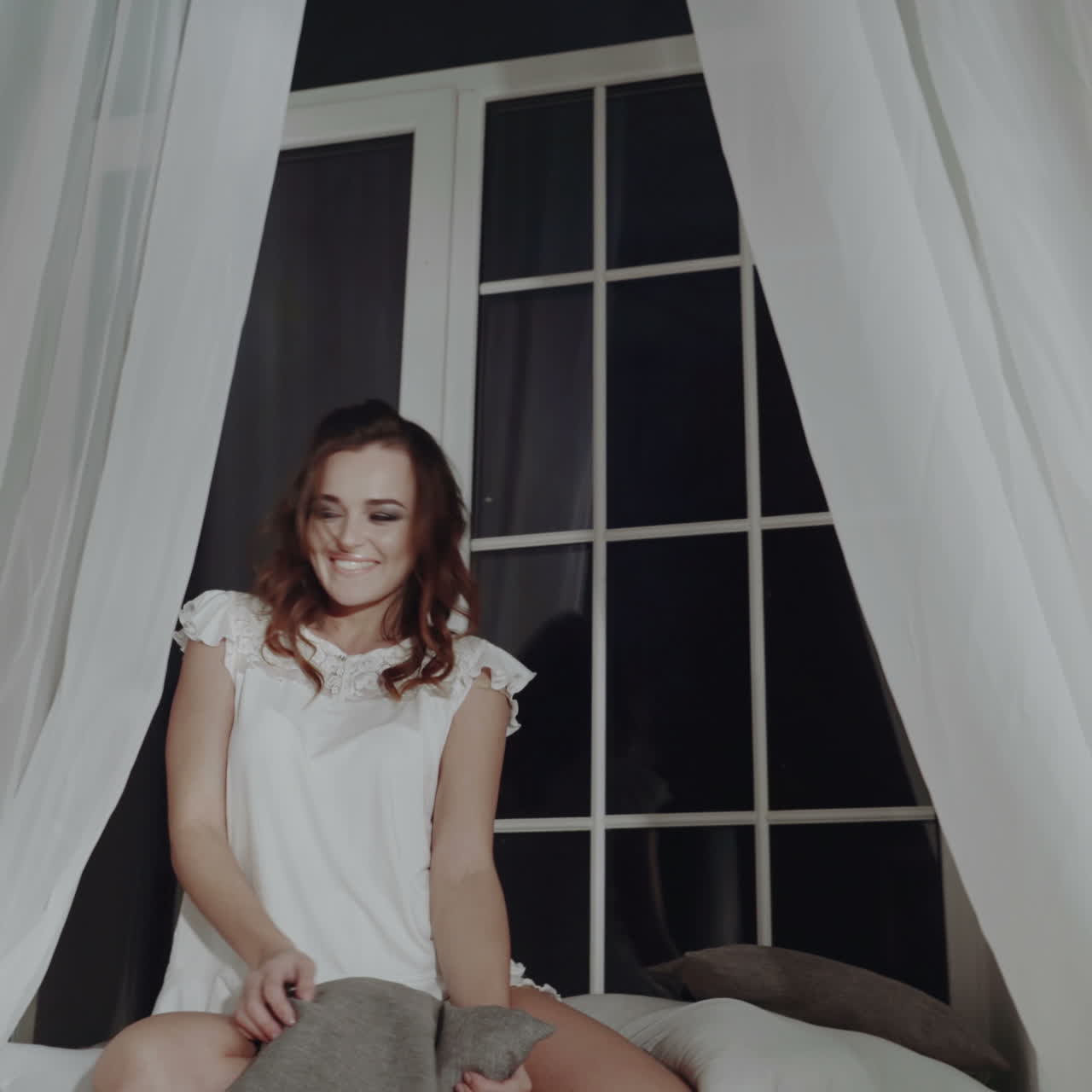 Woman in sleepwear, smiling, sitting on a bed by a window at night