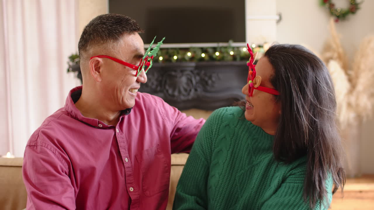 Senior asian couple laughing together wearing festive glasses at home during Christmas