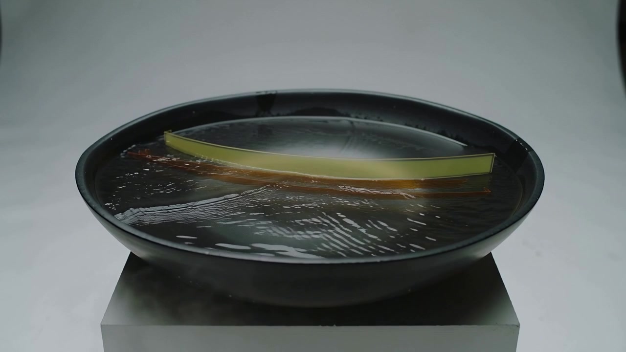 Rippling black bowl rocking yellow and amber strips from center ripple on pedestal forming waves