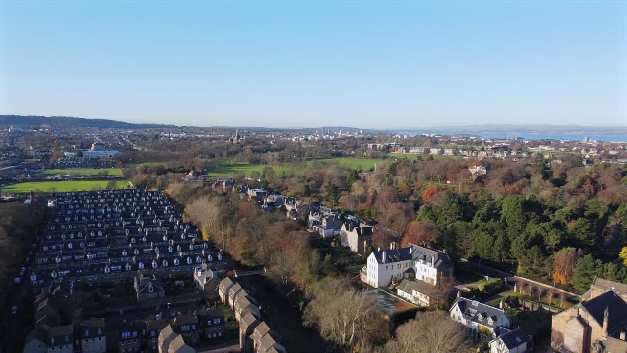 Droneoptagelser over et forstadskvarter i Edinburgh, Skotland, der viser rækker af huse, grønne områder og fjerne udsigter over byen og kystlinjen. Optagelserne fremhæver efterårstræerne