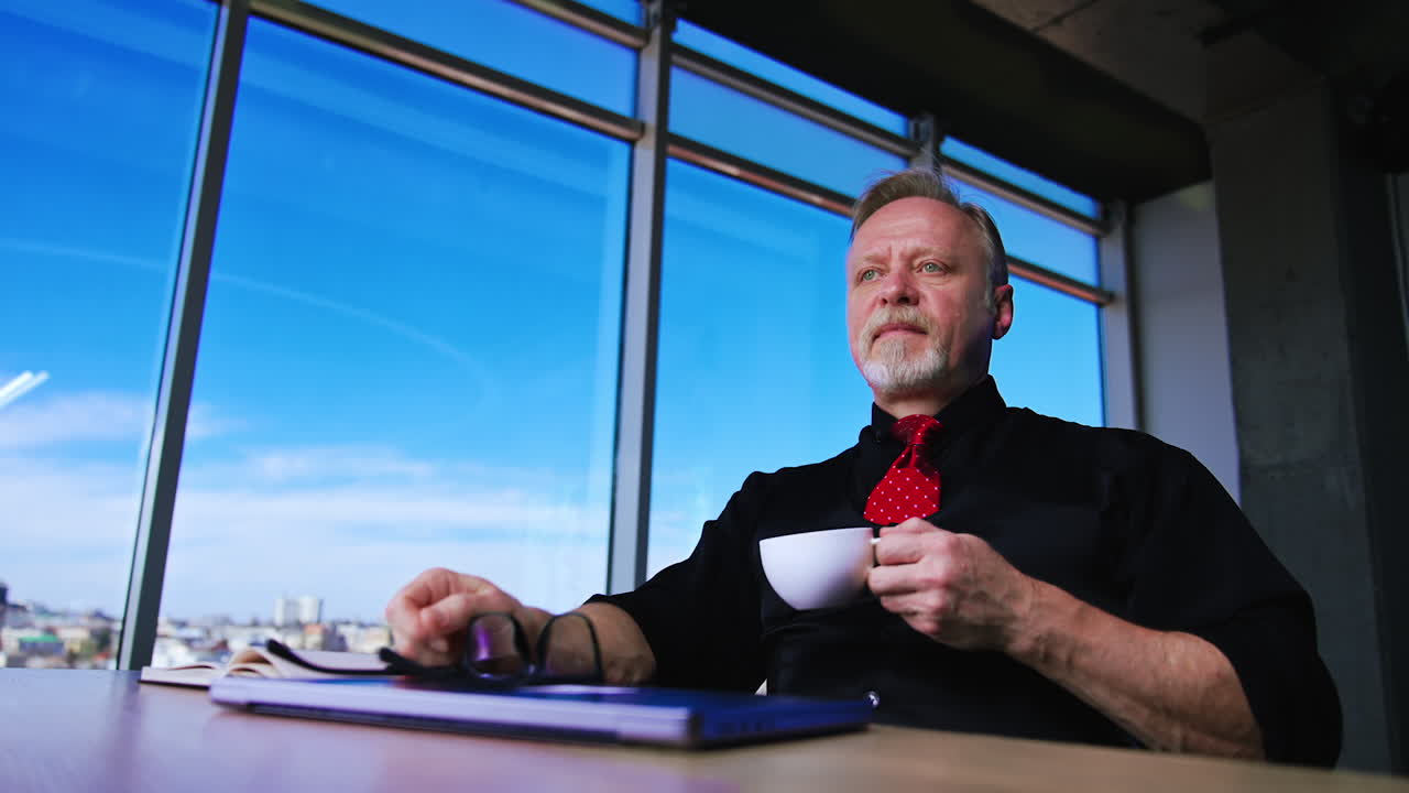 Calm confident Caucasian mature businessman sits at desk in office with cup in hand. Man takes off his glasses, puts them on laptop and drinks coffee. Low angle view.