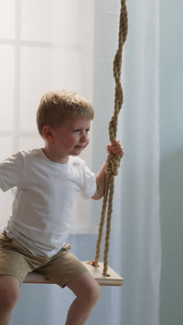 toddler boy exclaims and laughs contentedly on swing in cozy nursery with rack and light window. little son has fun at home. children entertainment