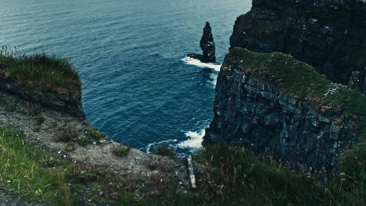 Low angle view of the Cliffs of Moher with blue sea, crashing waves, foreground rock and coastal greenery - Ireland