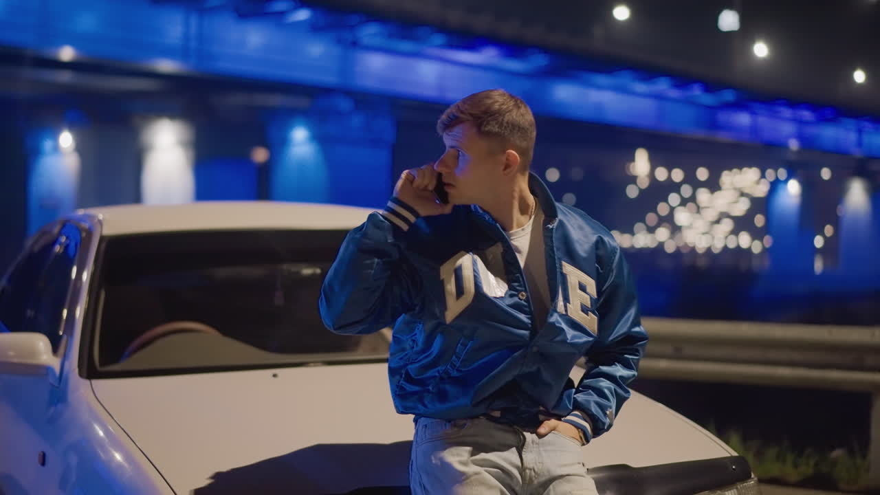 hombre en el capó del coche hablando por teléfono bajo las luces de un puente azul, apoyado en un sedán blanco con una chaqueta de satén, mirando hacia el agua, ambiente cinematográfico reflectante, carretera vacía, soledad nocturna