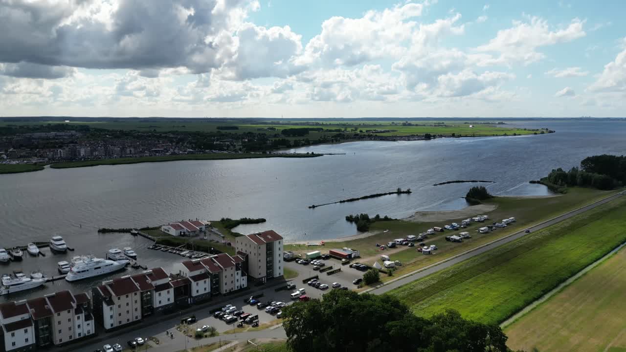 Charming riverside town with a marina, modern buildings, and lush green fields under a partly cloudy sky.