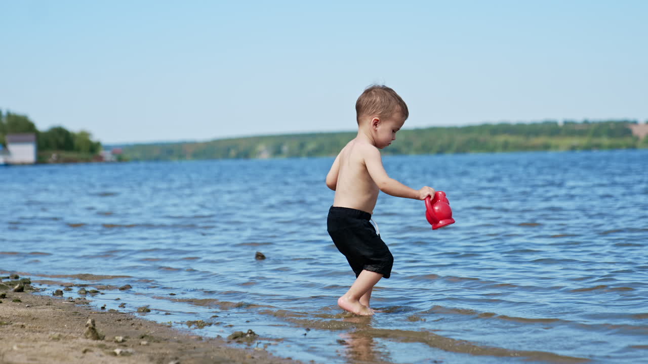 Lovely baby boy spending time at the beach in summer. Toddler walks into the water to fill his watering can.