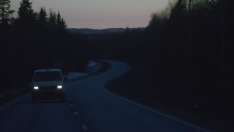 a car drives along an empty road at dusk through a forested landscape with its headlights on