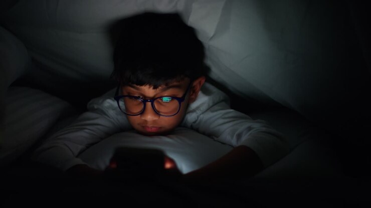 Young Boy In Bed Lying Under Duvet At Home At Night Looking At Mobile Phone With Motion Graphics Emojis Showing Multiple Social Media Or Messaging Notifications Liking And Reacting To Online Content