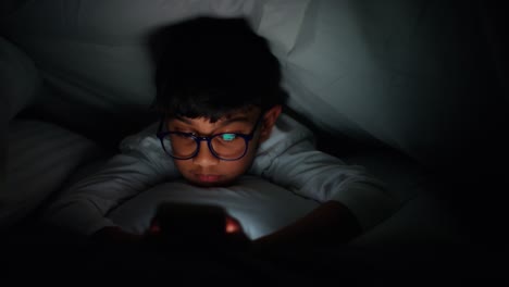 young boy in bed lying under duvet at home at night looking at mobile phone with motion graphics emojis showing multiple social media or messaging notifications liking and reacting to online content