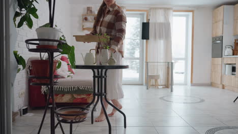 house keeper barefoot walks into cozy modern room carrying wooden tray sets tray on round table removes napkin from tray next to potted plants sofa sheer curtains framing snowy view outside