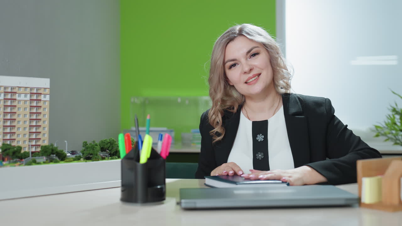 business mogul closes book while smiling at desk in brightly lit office with architectural model, laptop, pens, and greenery in view, conveying confidence, success, and professional atmosphere