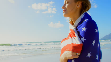 Seitenansicht-Einer-Jungen-Kaukasischen-Frau,-Die-In-Eine-Amerikanische-Flagge-Gehüllt-Ist-Und-Am-Strand-Spaziert,-4k