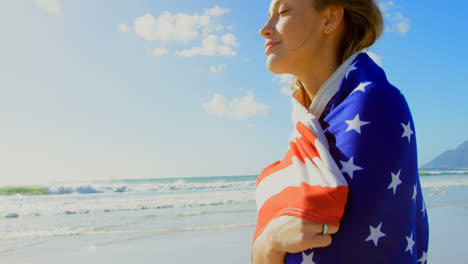 Vista-Lateral-De-Una-Joven-Caucásica-Envuelta-En-Una-Bandera-Americana-Caminando-Por-La-Playa-4k