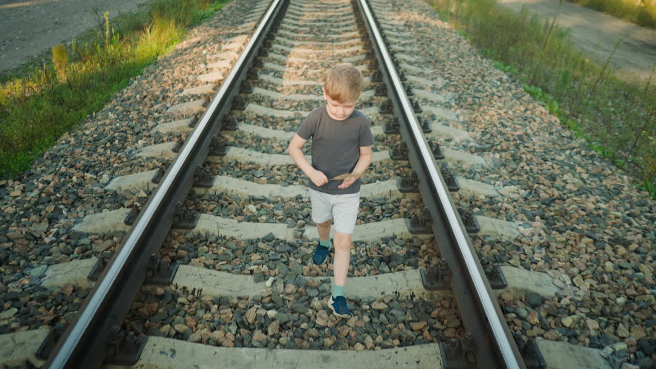 Young boy in casual outfit walking along rail track while holding dry grass in his hand, surrounded by gravel and rural path with soft natural light illuminating peaceful outdoor scene