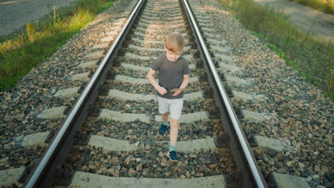 young boy in casual outfit walking along rail track while holding dry grass in his hand, surrounded by gravel and rural path with soft natural light illuminating peaceful outdoor scene