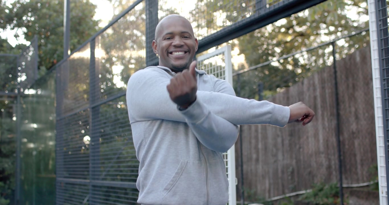 African American man stretching arm across chest while straddling court net, wearing hoodie