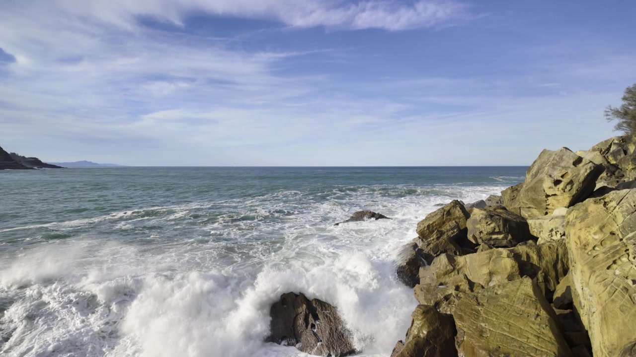 Grote krachtige oceaangolven die neerstorten op een rotsachtige kust in Getaria, Baskenland. Energiek wit schuim van de zee spat op de rotsen onder een prachtige blauwe lucht met enkele ijle wolken.