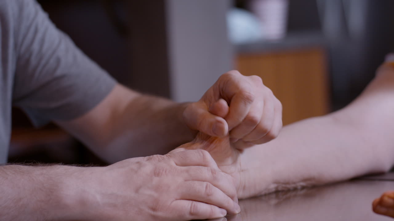 Close-up shot of middle-aged son massaging the hemiplegic right hand of senior mother stroke survivor, on top of a home glass top kitchen table. Focused on massaging thumb. 24fps.