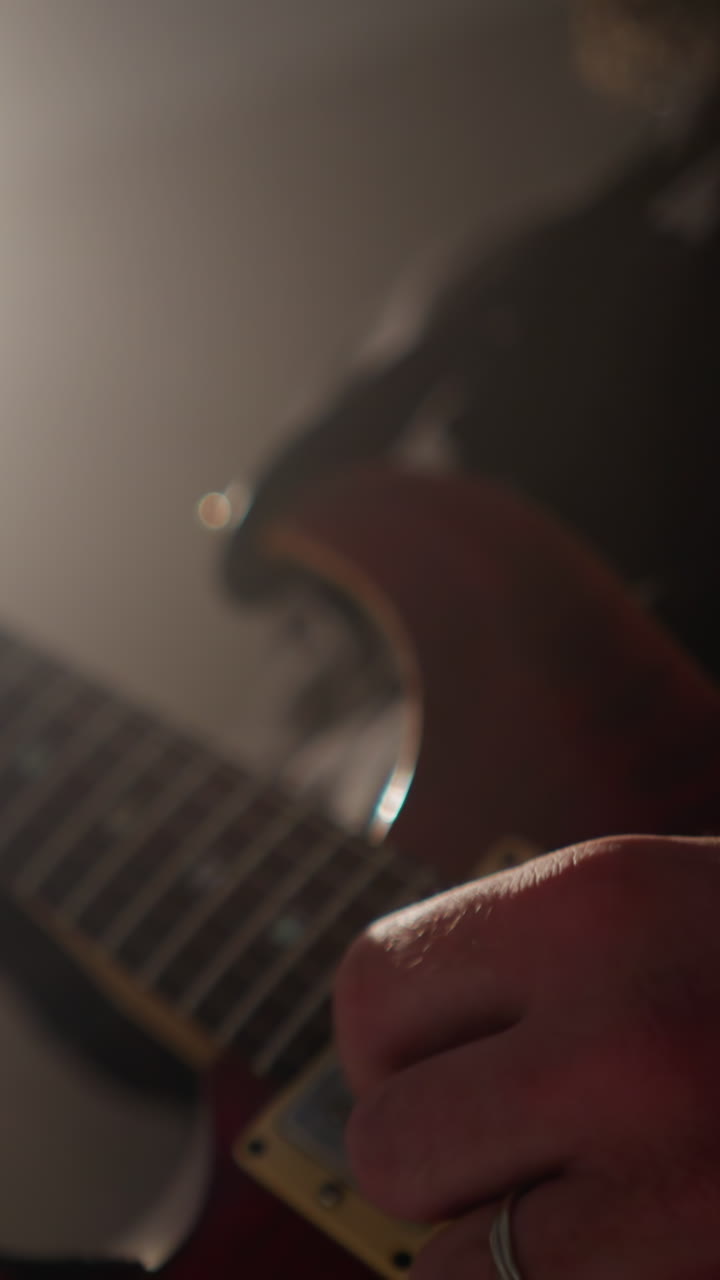 primer plano de las manos de un guitarrista tocando una guitarra roja, resaltado por un haz de luz enfocado en una habitación débilmente iluminada mientras la cámara hace zoom