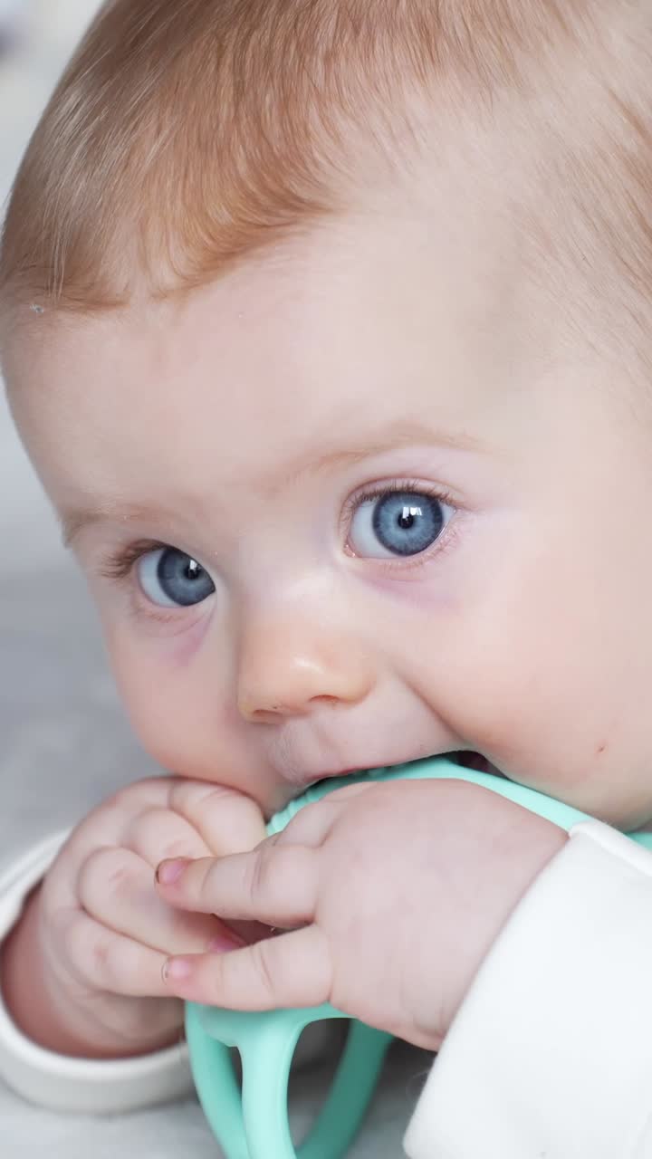 Vertical Close up portrait featuring a baby girl with striking blue eyes and soft light hair, happily chewing on a light blue teether, captured in a serene indoor setting in Riga, Latvia
