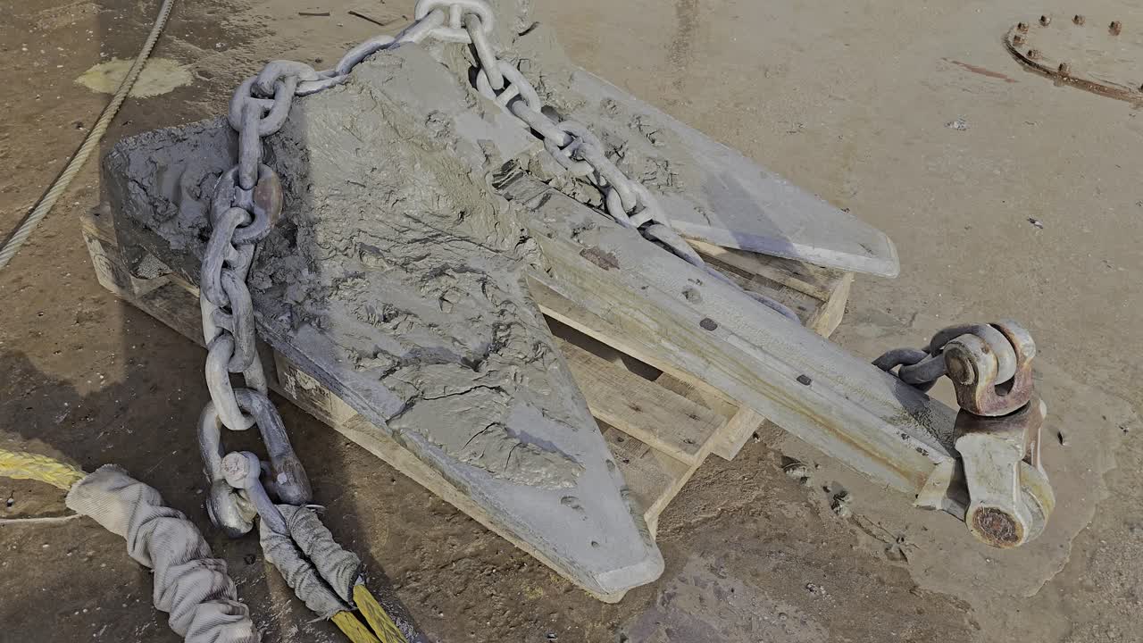 Dirty anchor rests on pallet in drydock. Mud and wear suggest recent use before inspection or repair