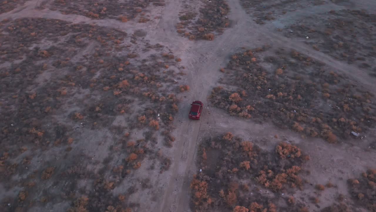 Top-down drone of a red SUV driving along a dusty desert track through sparse scrub; wide barren terrain, soft evening light—great for travel, off-road, and adventure B-roll