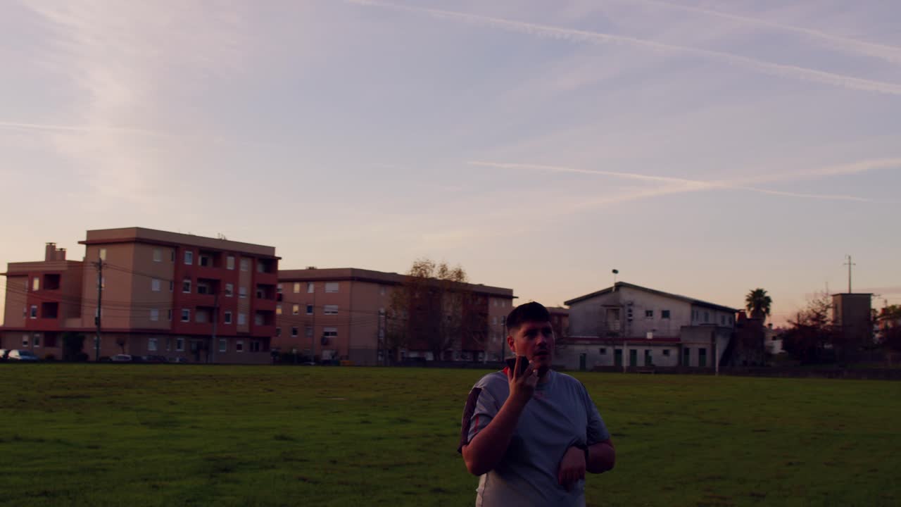 Young man in sportswear answering a call on a green field at dusk.