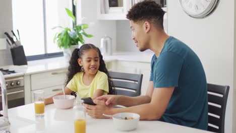 Glücklicher-Gemischtrassiger-Vater-Und-Tochter,-Die-Frühstücken-Und-Ihr-Smartphone-In-Der-Küche-Benutzen