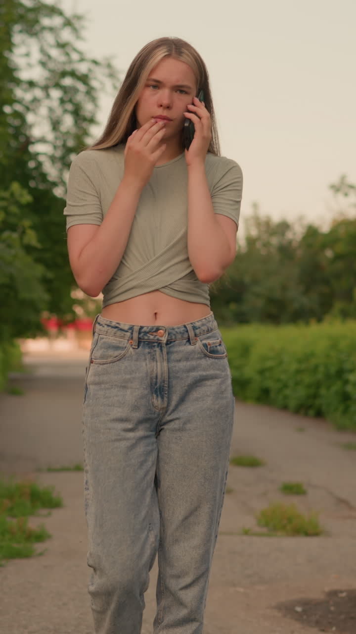mujer joven en vaqueros casuales y crop top gris caminando a lo largo de un camino bordeado de exuberante vegetación y árboles, involucrada en una conversación telefónica reflexiva con una mano cerca de su boca