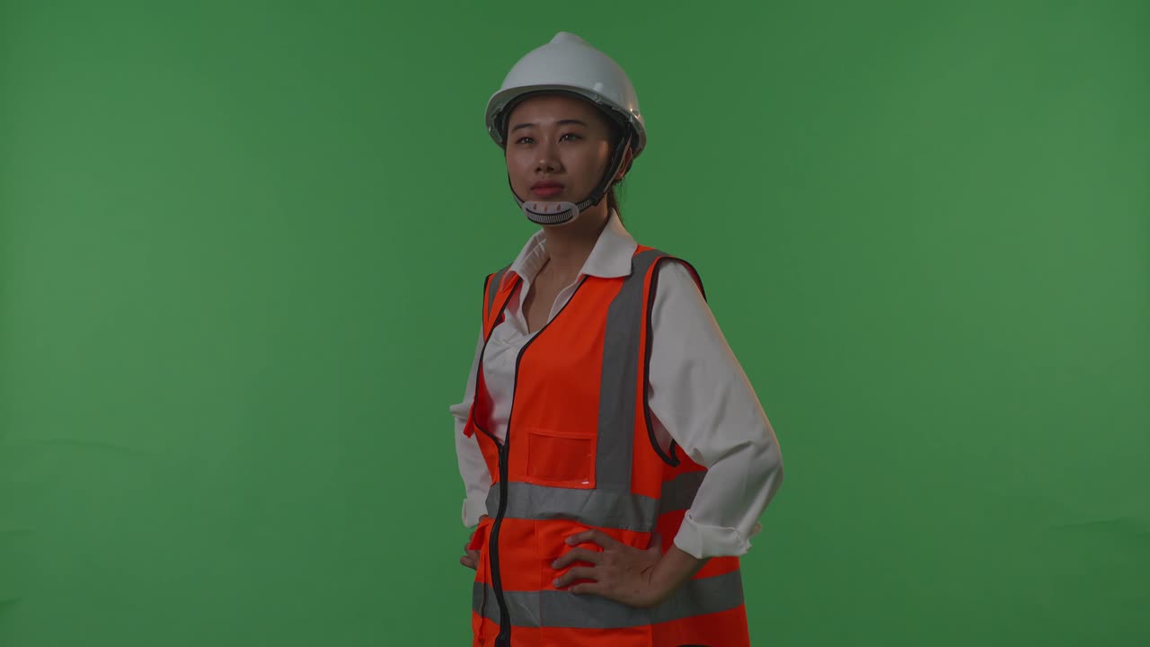 vista lateral de una ingeniera asiática con casco de seguridad mirando a su alrededor mientras está de pie con los brazos akimbo en el estudio de fondo de pantalla verde, durante la puesta o el amanecer