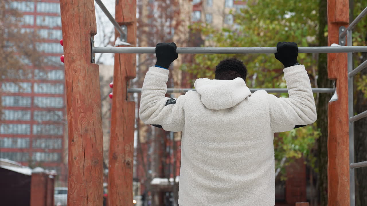 색 겨울 재을 입은 젊은 남자가 야외 체조 장비에 접근하고, 가을 설정에서 풀업을 위해 바를 잡기 위해 빠르게 점프합니다.