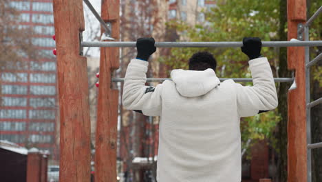 vista trasera de un joven con una chaqueta blanca de invierno que se acerca al equipo de gimnasia al aire libre, saltando rápidamente para agarrar la barra para un pull-up en un entorno de otoño