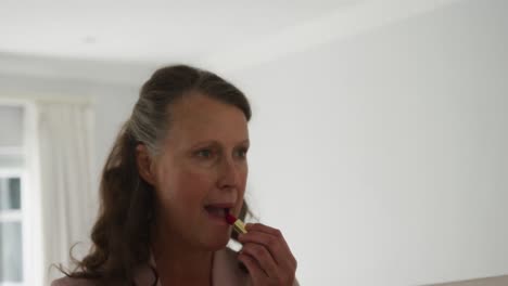 senior caucasian woman standing in room looking in mirror and putting on lipstick