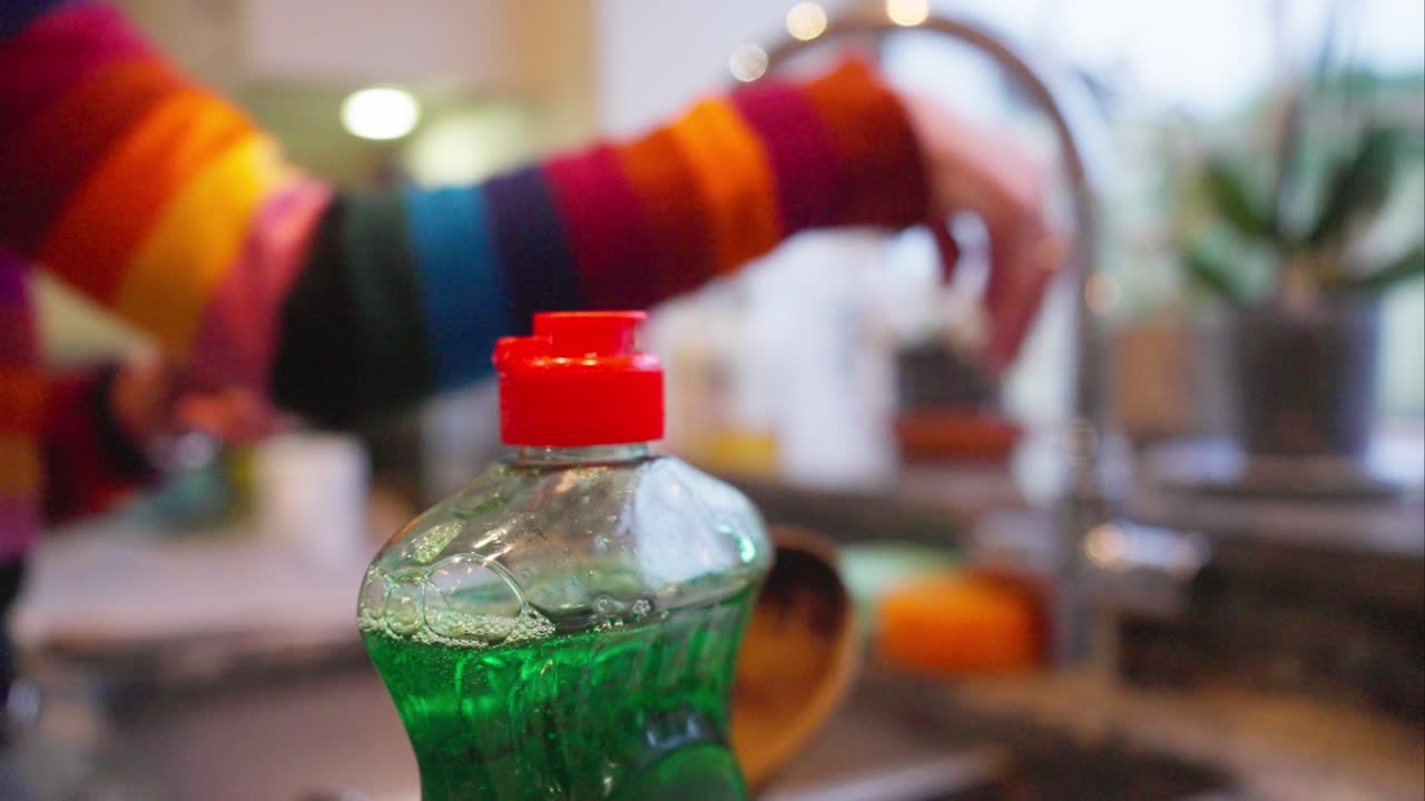 Focused washing up liquid in foreground, woman releases dirty water, blur sink