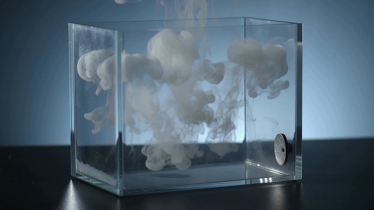 Entering from above white plume sinking, diffusing inside glass tank on dark table, metallic disc