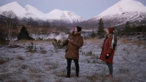 a caucasian couple launches drone at the field in front the mountains with snowy peaks on the background. man operating the copter, woman standing close. snowy countryside around