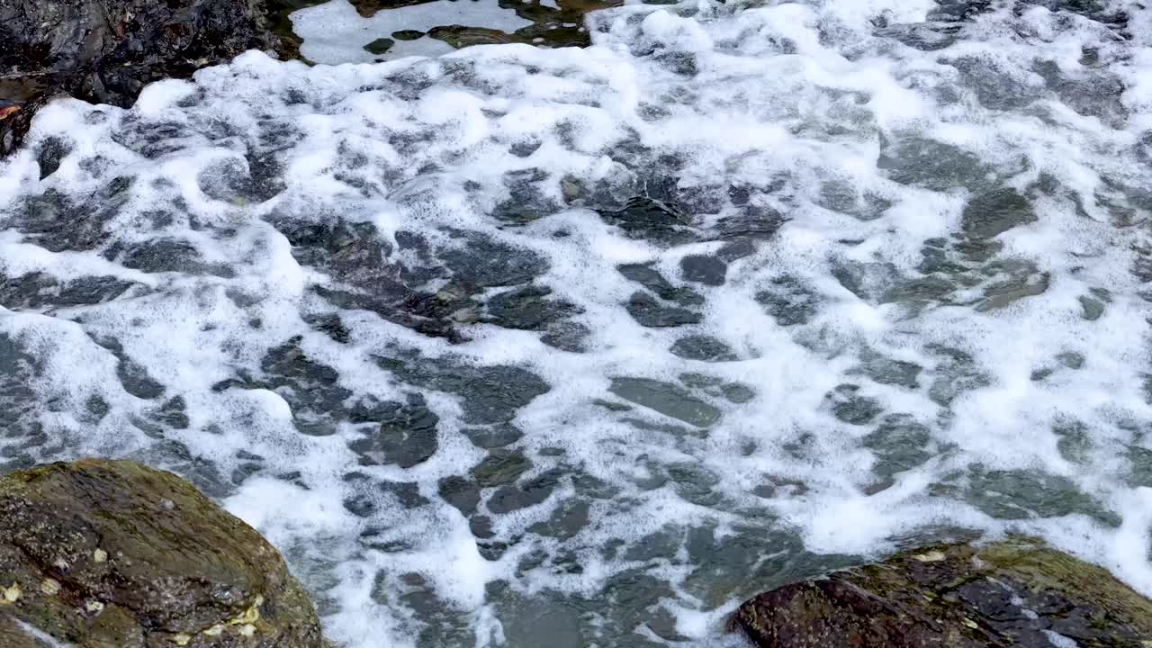 Skummende havbølger skyller ind over mørke kystklipper, dagslys, roligt kamera, naturligt havmiljø