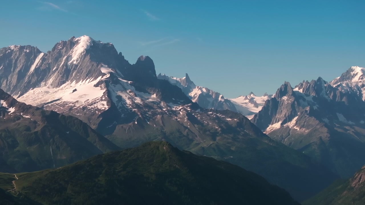 Rekaman udara puncak Alpen yang dramatis dan tertutup salju dengan punggung bukit bergerigi serta bercak gletser berlatar langit biru cerah
