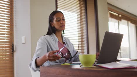 Frau-Mit-Gemischter-Abstammung,-Die-Einen-Laptop-Benutzt-Und-In-Ein-Notizbuch-Schreibt,-Kaffee-Trinkt-Und-Von-Zu-Hause-Aus-Arbeitet