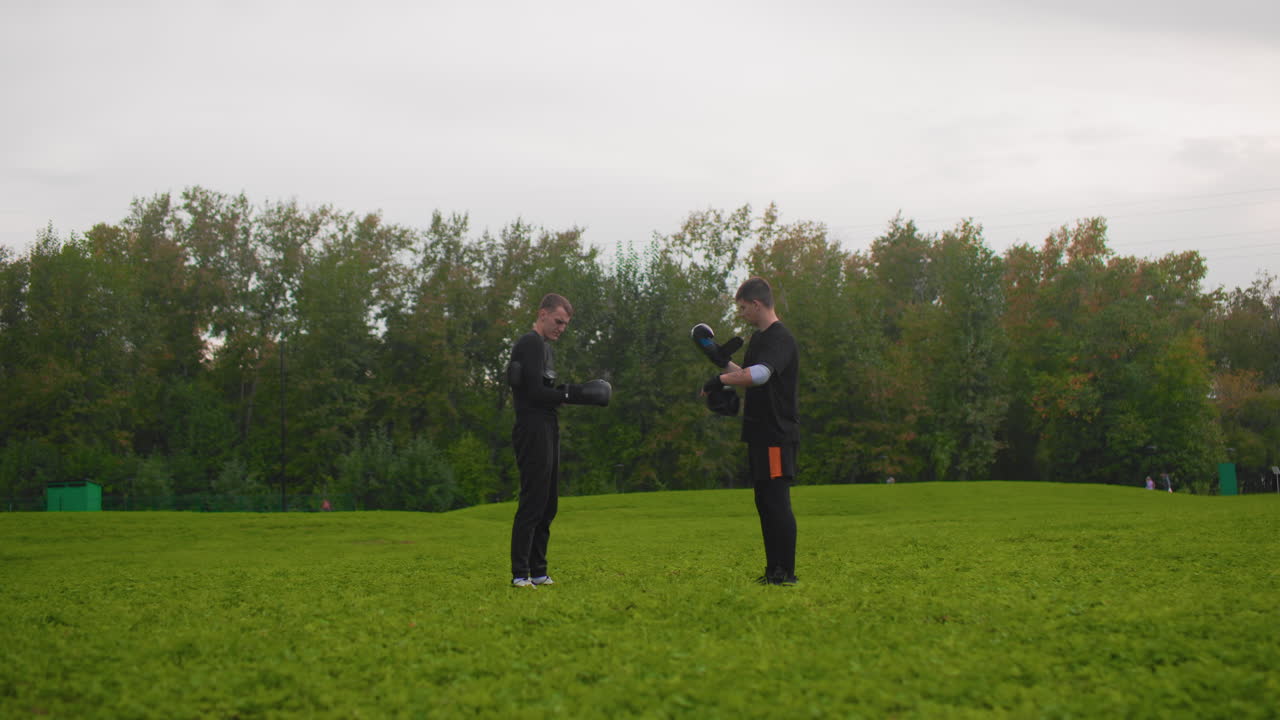 In un parco aperto, due uomini bianchi si stringono la mano e si scambiano suggerimenti di addestramento, regolano i guanti e si danno brevi istruzioni, un duo che pratica la postura e il gioco di gambe su un prato verde con alberi sullo sfondo, in un'atmosfera rilassata e di supporto.