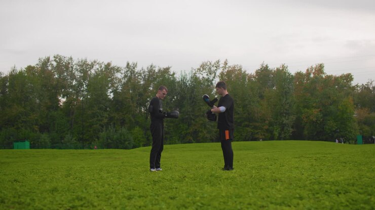 In un parco aperto, due uomini bianchi si stringono la mano e si scambiano suggerimenti di addestramento, regolano i guanti e si danno brevi istruzioni, un duo che pratica la postura e il gioco di gambe su un prato verde con alberi sullo sfondo, in un'atmosfera rilassata e di supporto.