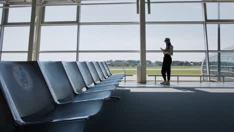 woman traveler, leisure or business standing at the glass window of a large international airport hub with a phone in his hands, scrolling the application for smartphones. in the hands of tickets