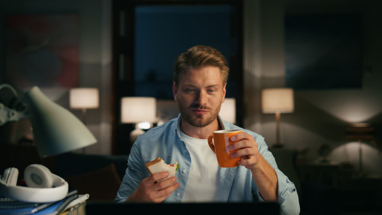 Positiv mand videoopkalder hjemmefra, nærbillede. Fyr drikker og tager en sandwichpause.