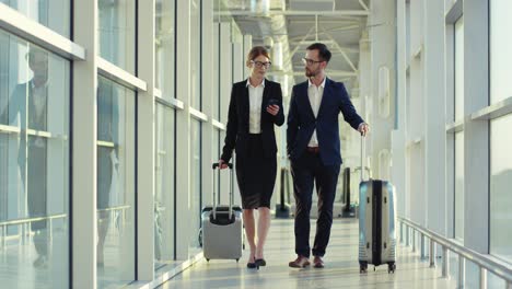 attractive man and woman in business style walking the corridor of the airport with suitcases, speaking and woman tapping on the smartphone