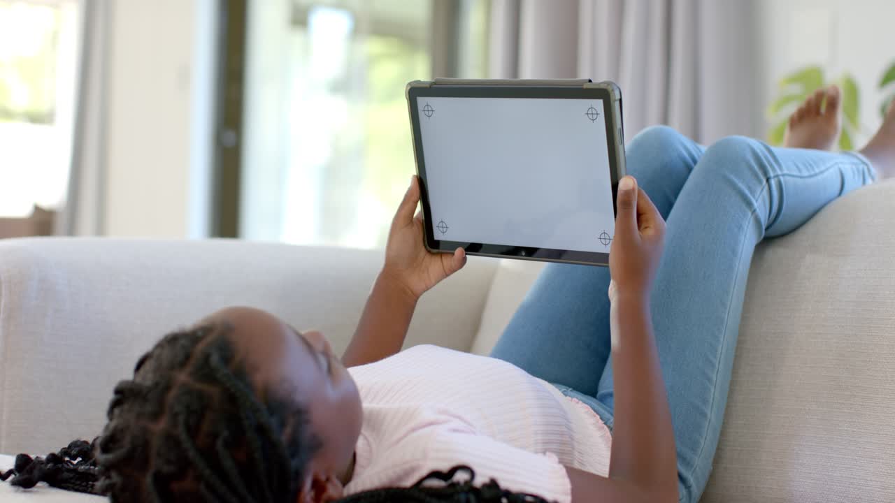 Clip starting showing child reclining on sofa near window holding tablet, shifting grip for comfort