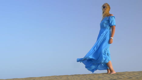 slow motion view of young blond woman standing against blue sky in long blue dress and it is fluttering on the wind